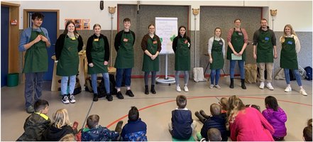 Studierende stehen in der Kita Purzelbaum in einer Reihe und tragen grüne Schürzen. Im Vordergrund sitzt, ihnen zugewandt, eine Gruppe von Kindern.rund sitzen Kinder.