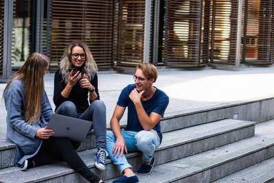 Studierende sitzen vor einem Universitätsgebäude auf Treppenstufen.