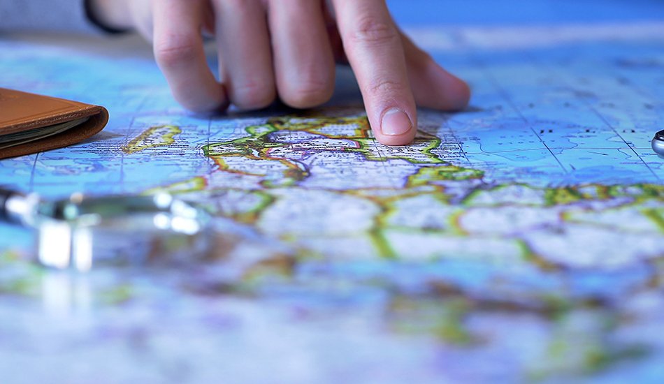 Referat Internationales (motortion/AdobeStock) Finger auf Weltkarte. Im Vordergrund liegt eine Lupe.