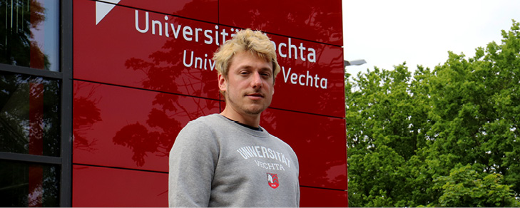 Student im grauen Uni Vechta Pullover vor Roter Wand.
